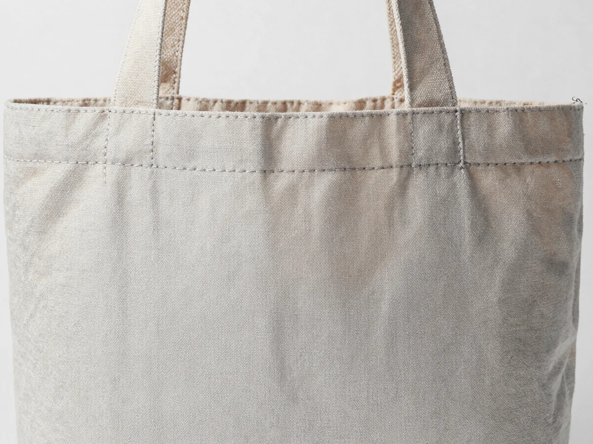 Macro close-up of tight, neat stitching on a natural canvas tote bag, soft light-grey background, highlighting texture.